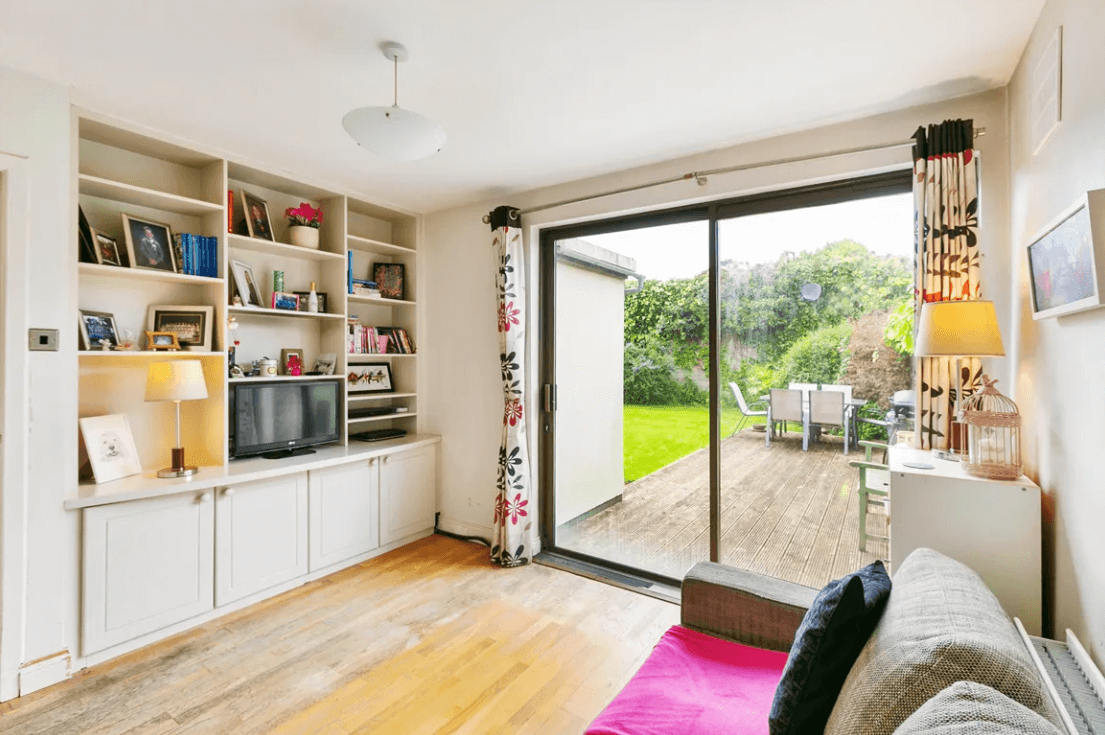 129 Wesbury Stillorgan Dublin Family Room with white walls and ceiling, timber flooring, large sliding doors to the rear garden, floor-to-ceiling bookcases and white storage units and a coffee-coloured sofa.