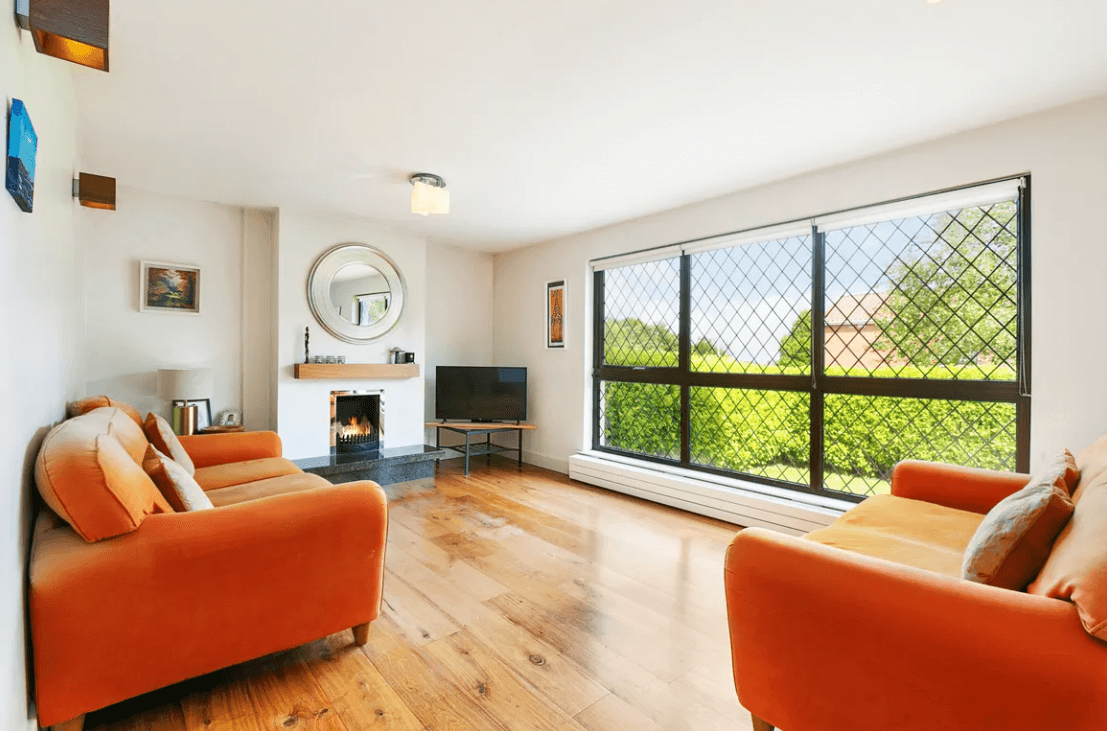 Living Room with white walls and ceiling, timber flooring, large lattice-style window to the front garden, fireplace with lare oval mirror above, TV stand in the corner and an orange-coloured sofa.