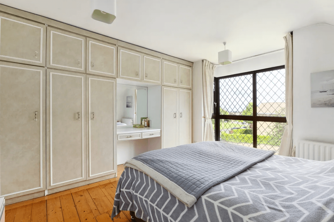 ublin Principal bedroom with white ceiling, large bed, timber flooring, large bed, white fitted wardrobes, and medium-sized window with view of garden