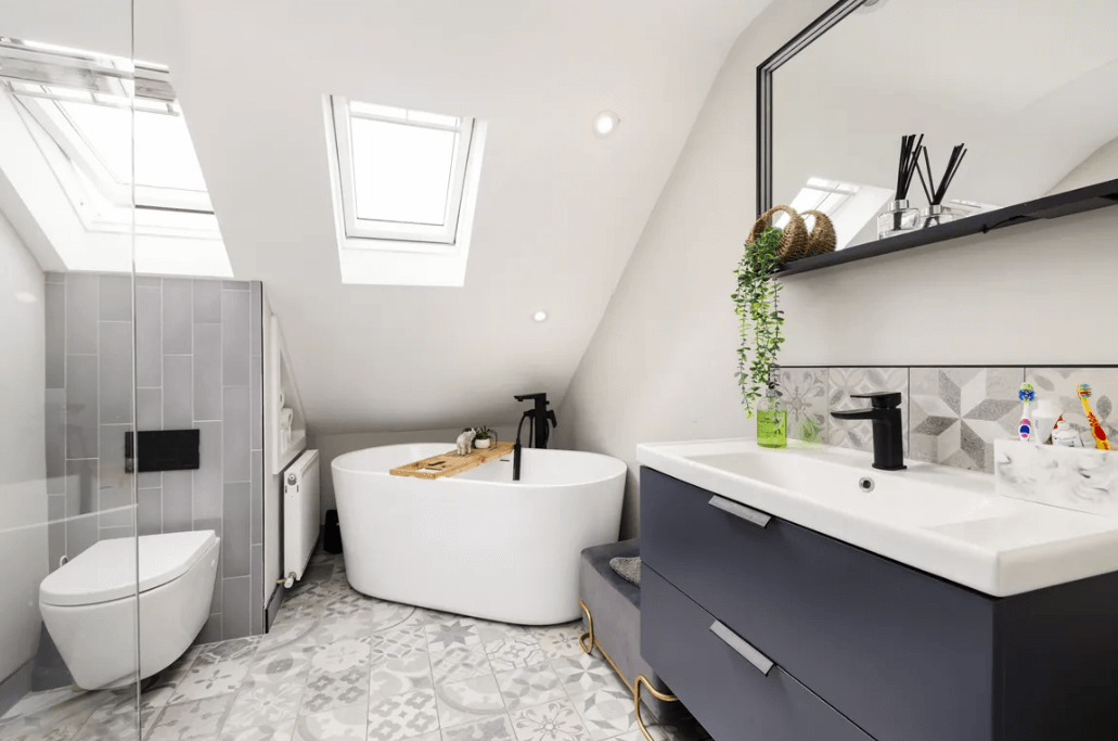 17 The Courtyard Brookwood Bray bathroom in a very modern style with independent white bath, grey tiled flooring, shower unit and sink area with mirror and Velux window in the roof