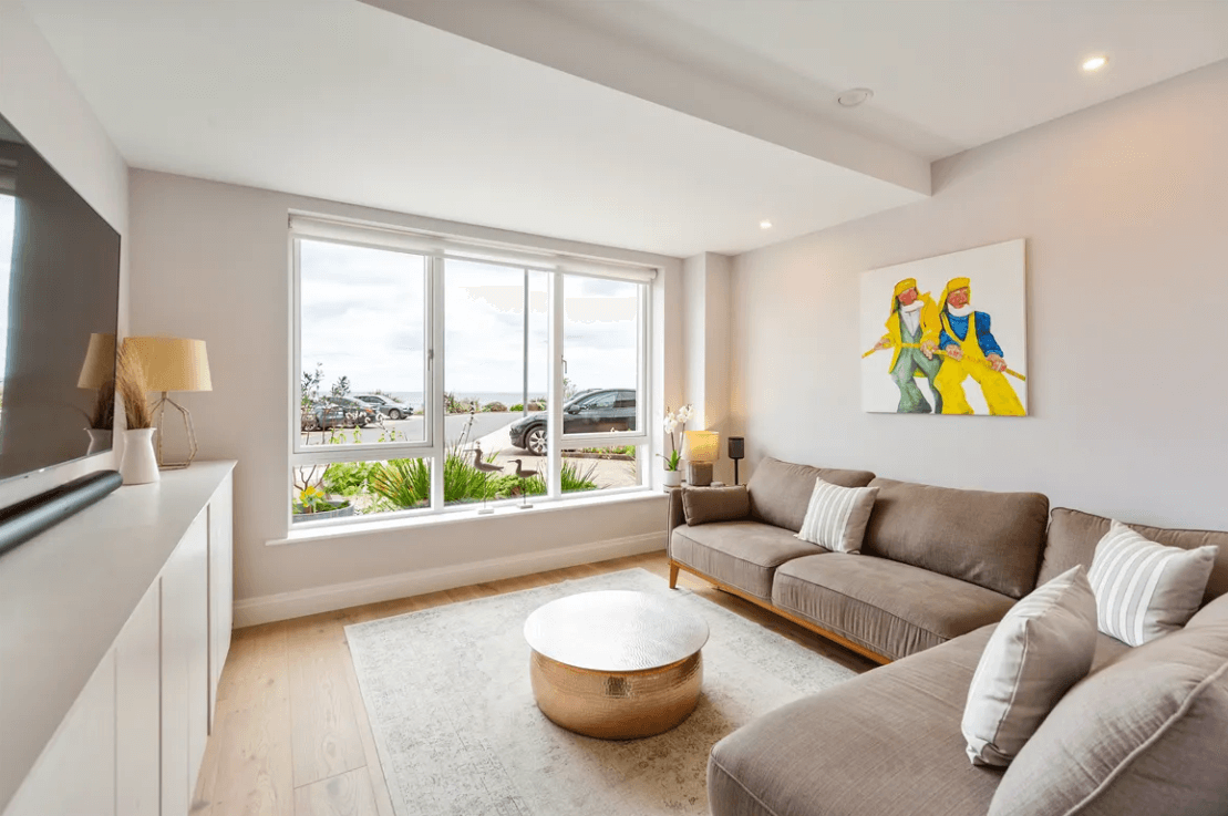 16 The Shore Greystones Wicklow George and Maguire Properties living room with carpet rug on a timber floor with L-shaped light brown sofa and large window with a front view